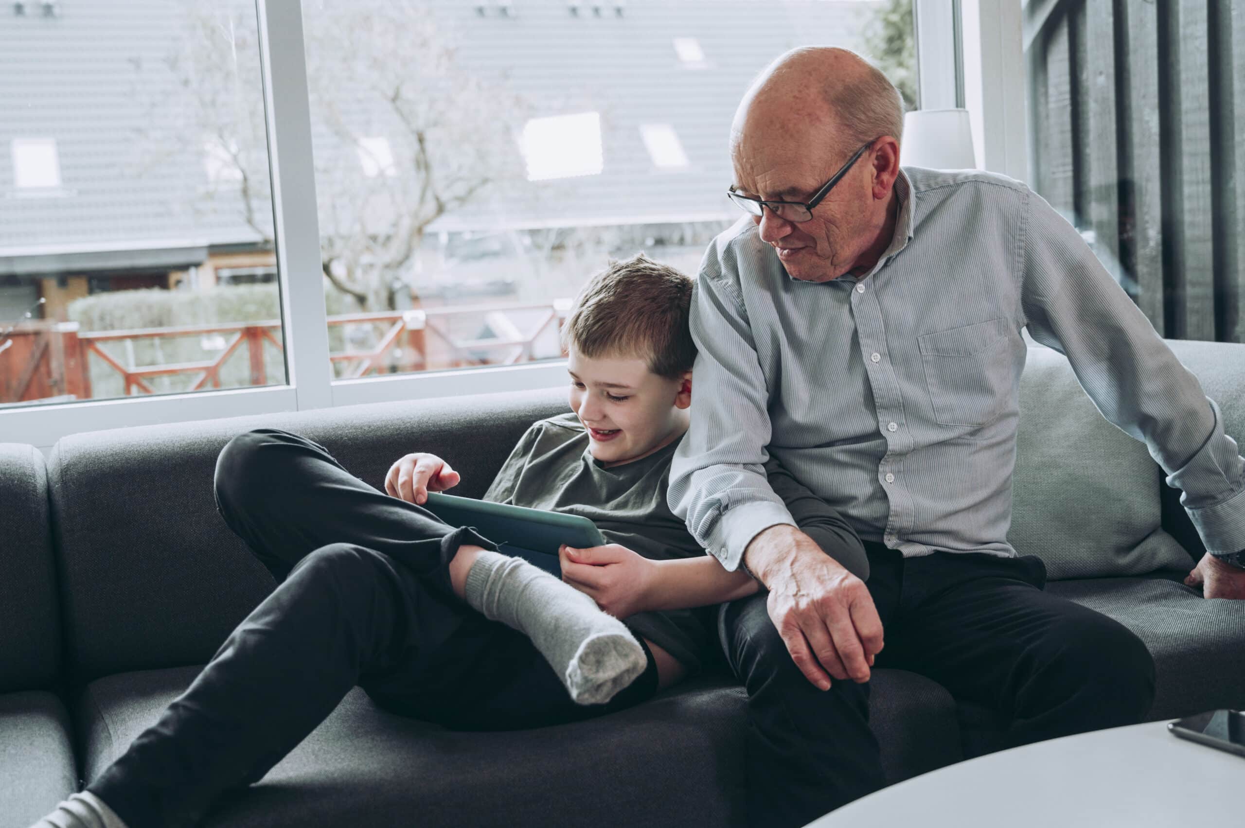Grandfather and grandson enjoy spending time together playing with tablet at home Grandfather and grandchild sit in a sofa in a living room. The boy holds on to a digital tablet, and they both look at the screen.